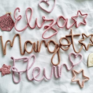May include: Crochet name signs and shapes in pink and brown. The names "Eliza", "Margot", and "Fleur" are displayed, along with star and heart shapes. A small wooden sign says "Stay Wild".