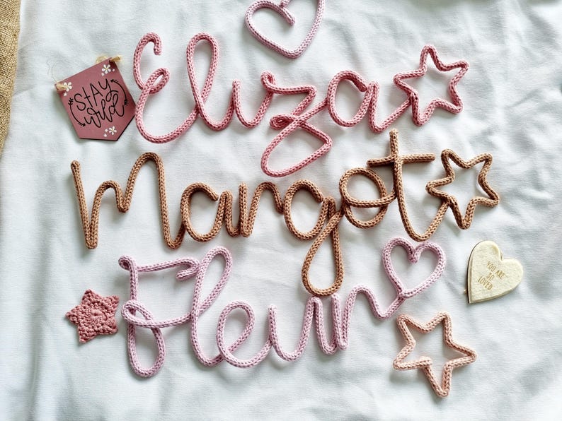 May include: Crochet name signs and shapes in pink and brown. The names "Eliza", "Margot", and "Fleur" are displayed, along with star and heart shapes. A small wooden sign says "Stay Wild".