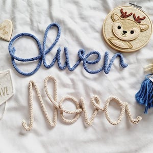 May include: A selection of nursery decorations, featuring blue and cream yarn name signs spelling "Oliver" and "Nova". Also includes a heart tag, a felt banner with "BE BRAVE", a deer embroidery hoop, and a small doll.