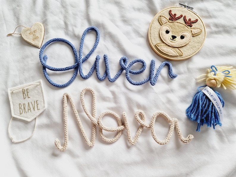 May include: A selection of nursery decorations, featuring blue and cream yarn name signs spelling "Oliver" and "Nova". Also includes a heart tag, a felt banner with "BE BRAVE", a deer embroidery hoop, and a small doll.