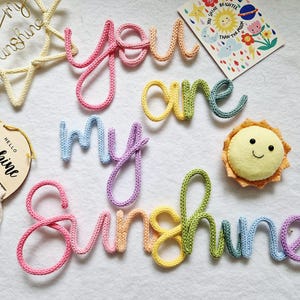 May include: A selection of handmade decorations with a sunshine theme. Features a wire sun, a wooden plaque with macrame, a felt sun, and the phrase "you are my sunshine" made from colourful yarn.