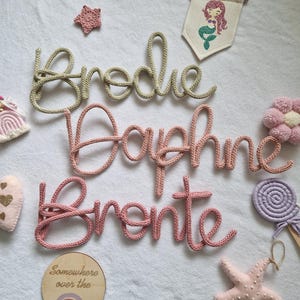 May include: A selection of nursery decorations featuring three knitted name signs: "Brodie", "Daphne", and "Bronte", in a cursive style. The image also includes a mermaid banner, a star, a wooden sign with a rainbow, and other decorative items.