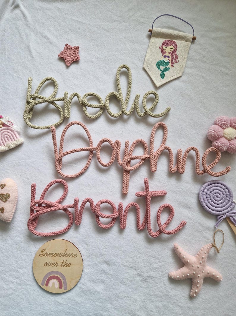 May include: A selection of nursery decorations featuring three knitted name signs: "Brodie", "Daphne", and "Bronte", in a cursive style. The image also includes a mermaid banner, a star, a wooden sign with a rainbow, and other decorative items.