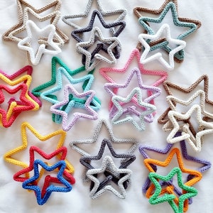 May include: A collection of 12 crocheted star decorations in various colours, including red, yellow, orange, green, blue, purple, white, and brown. The stars are layered on top of each other, creating a visually appealing display.