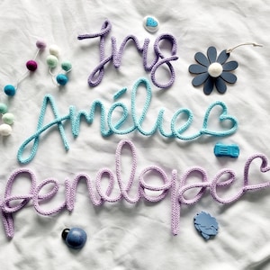 May include: Knitted name signs in purple, turquoise, and lilac. The names "Iris", "Am&eacute;lie", and "Penelope" are displayed in cursive script. Decorative elements include a felt ball garland, a flower, and small embellishments.