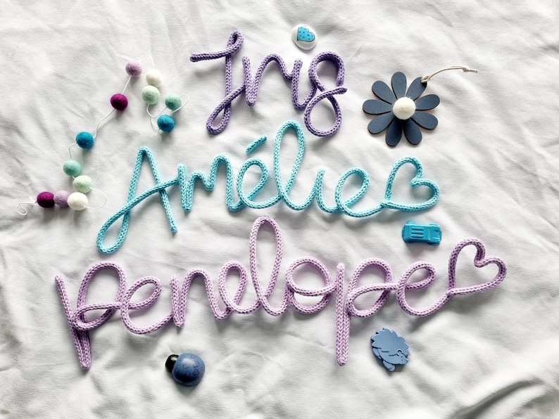 May include: Knitted name signs in purple, turquoise, and lilac. The names "Iris", "Am&eacute;lie", and "Penelope" are displayed in cursive script. Decorative elements include a felt ball garland, a flower, and small embellishments.