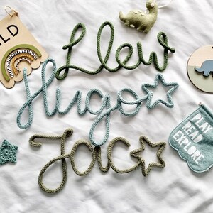 May include: A selection of nursery decorations, featuring name signs and a flag with the words "PLAY READ EXPLORE". The names "Llew", "Hugo", and "Jac" are crafted from green and blue yarn. A wooden sign displays the word "WILD".