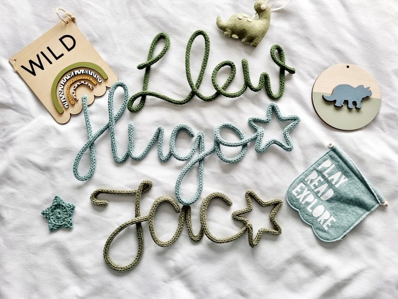 May include: A selection of nursery decorations, featuring name signs and a flag with the words "PLAY READ EXPLORE". The names "Llew", "Hugo", and "Jac" are crafted from green and blue yarn. A wooden sign displays the word "WILD".