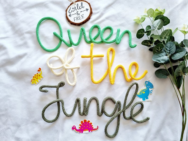 May include: Green, yellow, and white yarn letters spelling "Queen of the Jungle" with a wooden sign that says "Wild & Free" and dinosaur stickers on a white background.