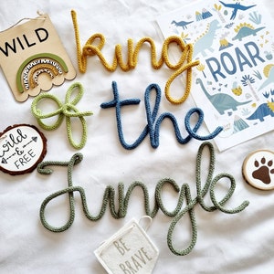 May include: A set of knitted word decorations spelling "king of the jungle" in green, yellow, and blue. There are also several wooden signs with the words "wild", "wild & free", and "be brave".