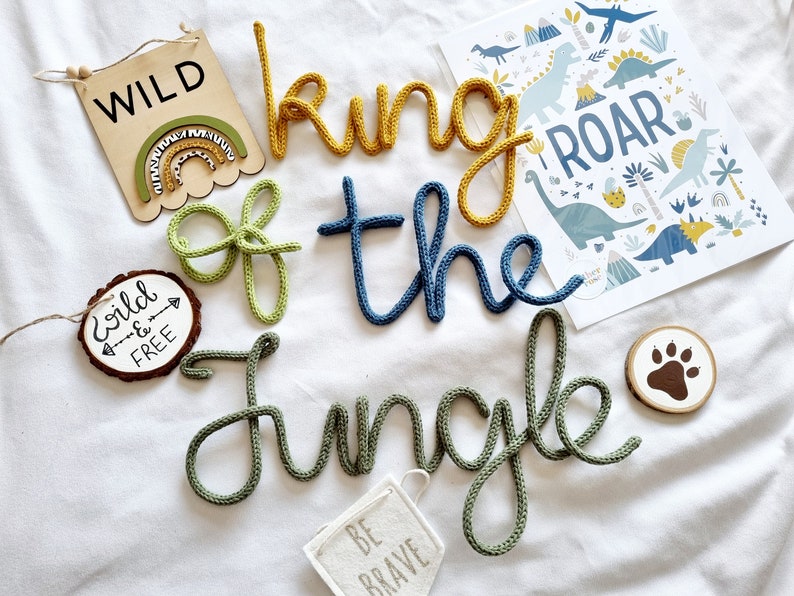 May include: A set of knitted word decorations spelling "king of the jungle" in green, yellow, and blue. There are also several wooden signs with the words "wild", "wild & free", and "be brave".