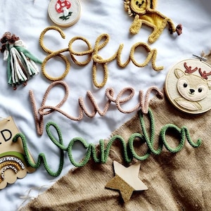 May include: A selection of handmade nursery decorations. The image features knitted name signs in yellow, pink, and green, a wooden sign with the word "WILD", a felt deer embroidery hoop, a lion, a mushroom, a star, and a doll.