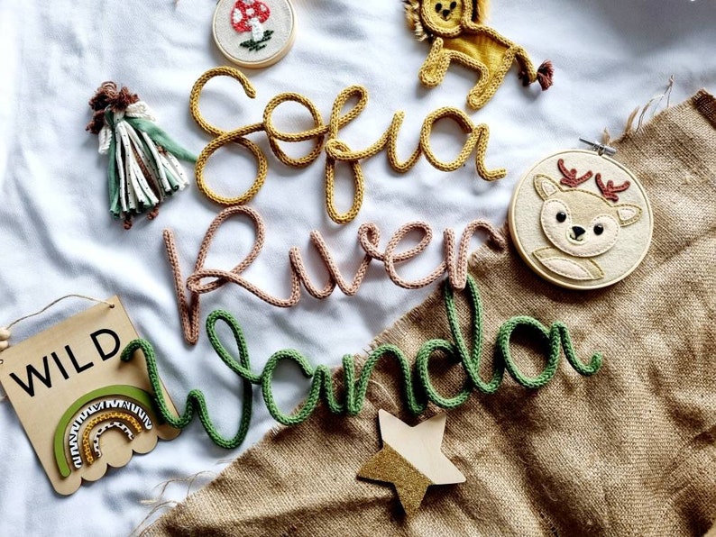 May include: A selection of handmade nursery decorations. The image features knitted name signs in yellow, pink, and green, a wooden sign with the word "WILD", a felt deer embroidery hoop, a lion, a mushroom, a star, and a doll.