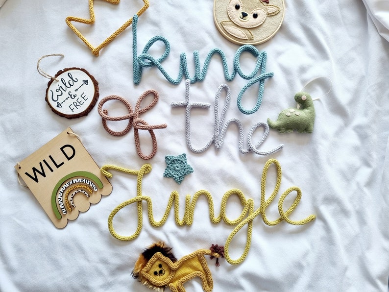 May include: A knitted nursery decoration with the words "King of the Jungle" in yellow, blue, and brown yarn. The decoration also includes a wooden sign with the word "WILD" and a rainbow, a wooden sign with the words "Wild & Free", a knitted star, a knitted deer, a knitted dinosaur, and a knitted lion.