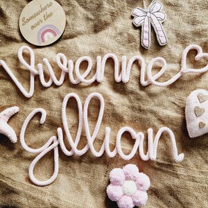May include: A light pink knitted name sign spelling "Vivienne Gillian" with a heart at the end of the first name. The sign is surrounded by decorative elements, including a starfish, a bow, a flower, and a wooden disc with a rainbow.
