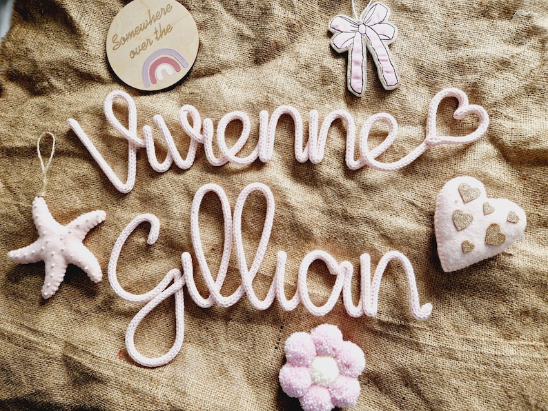 May include: A light pink knitted name sign spelling "Vivienne Gillian" with a heart at the end of the first name. The sign is surrounded by decorative elements, including a starfish, a bow, a flower, and a wooden disc with a rainbow.