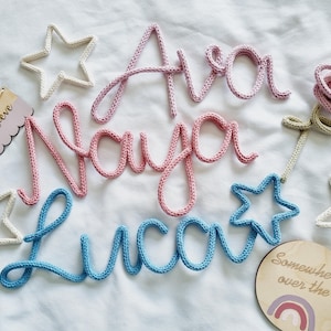 May include: A selection of nursery decorations, featuring the names "Ava", "Naya", and "Luca" in pink and blue yarn. The image includes star-shaped ornaments and a wooden sign with the word "Believe".
