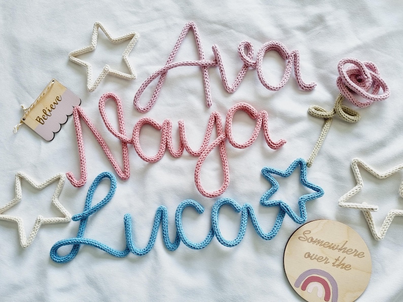 May include: A selection of nursery decorations, featuring the names "Ava", "Naya", and "Luca" in pink and blue yarn. The image includes star-shaped ornaments and a wooden sign with the word "Believe".