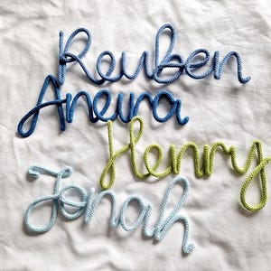 May include: Four names, Reuben, Aneura, Henry, and Jonah, made from blue and green yarn, are displayed on a white fabric. The names are in a cursive style, with each letter connected, creating a personalised and decorative element.