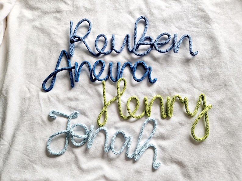 May include: Four names, Reuben, Aneura, Henry, and Jonah, made from blue and green yarn, are displayed on a white fabric. The names are in a cursive style, with each letter connected, creating a personalised and decorative element.