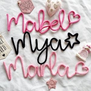 May include: Handmade yarn name signs in pink and black, spelling "Mabel", "Mya", and "Marla", with heart and star details. A small banner reads "You are loved", and a wooden sign says "Stay Wild". Decorative items are scattered on a white surface.