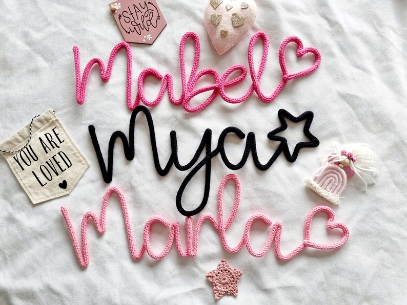 May include: Handmade yarn name signs in pink and black, spelling "Mabel", "Mya", and "Marla", with heart and star details. A small banner reads "You are loved", and a wooden sign says "Stay Wild". Decorative items are scattered on a white surface.