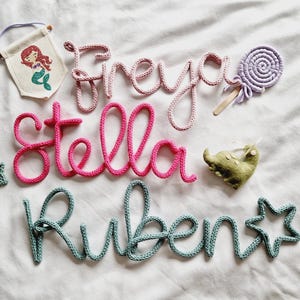May include: Handmade nursery decorations with the names "Freya", "Stella", and "Ruben" in pink and teal yarn. Includes a mermaid banner, a felt elephant, a star, and a lollipop.
