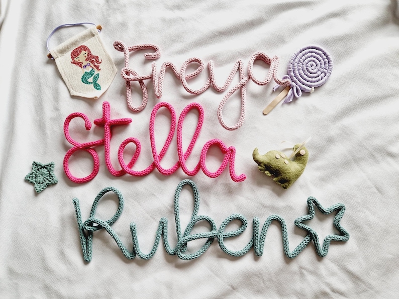 May include: Handmade nursery decorations with the names "Freya", "Stella", and "Ruben" in pink and teal yarn. Includes a mermaid banner, a felt elephant, a star, and a lollipop.