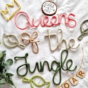 May include: A set of crocheted word art pieces spelling "Queens of the Jungle" in pink, beige, and green yarn. The words are decorated with small crocheted leaves. A wooden sign with the text "Wild & Free" is in the background.