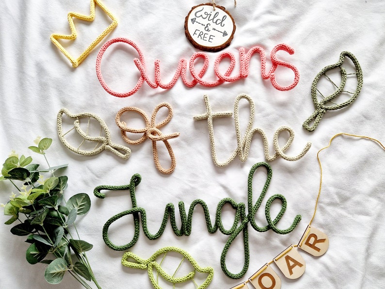 May include: A set of crocheted word art pieces spelling "Queens of the Jungle" in pink, beige, and green yarn. The words are decorated with small crocheted leaves. A wooden sign with the text "Wild & Free" is in the background.