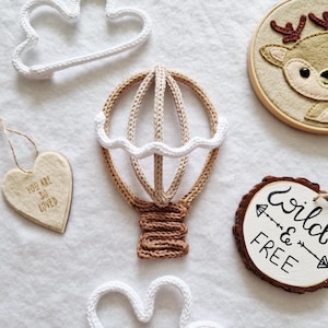Puede incluir: Una colección de artículos decorativos que incluye un globo aerostático tejido en beige y marrón, dos formas de nubes tejidas blancas, un adorno de fieltro en forma de corazón con el texto YOU ARE SO LOVED, un bastidor de bordado de ciervo y un disco de madera con el texto WILD & FREE.