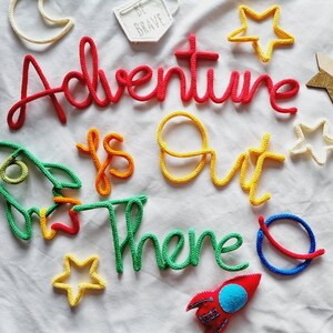 May include: A white background with colourful yarn letters spelling "Adventure is out there". There are also yarn stars, a rocket, a moon, and a small sign that says "Be Brave".