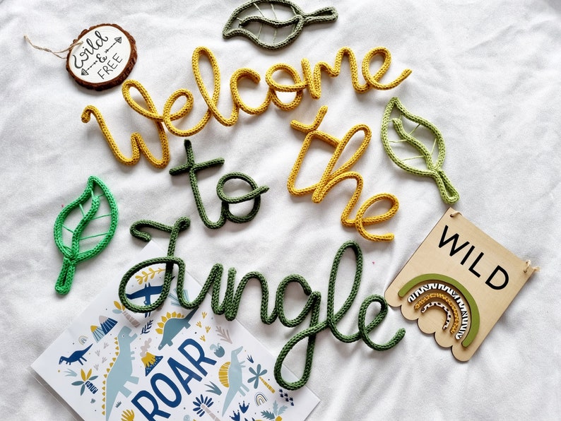 May include: Welcome to the jungle! A set of crocheted letters spelling out "Welcome to the Jungle" in green and yellow yarn. There are also two crocheted green leaves and a wooden sign that says "WILD" with a rainbow design.