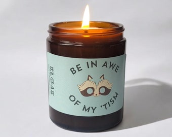 Pode incluir: Uma vela de vidro marrom com um rótulo azul claro que diz "BE IN AWE OF MY 'TISM" com uma ilustração de guaxinim de desenho animado. A vela está acesa e a chama é visível.
