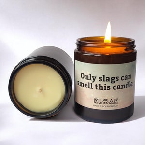 Puede incluir: Dos velas en frasco de vidrio &aacute;mbar con cera color crema. Una vela est&aacute; encendida con una llama visible. La etiqueta presenta un fondo degradado con estrellas y el texto: Only slags can smell this candle. KLOAK MADE IN BOURNEMOUTH.