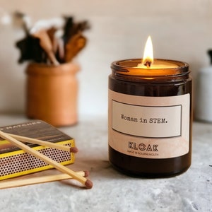 May include: A brown glass candle with a white label that reads "Woman in STEM." The candle is lit and has a flame. The candle is on a grey surface with a box of matches and an orange.