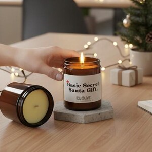 May include: A lit brown glass candle with a white label that reads "Basic Secret Santa Gift." Another candle is next to it. A small Christmas tree, a mug, and a notebook are on the table. A person is writing in the notebook.