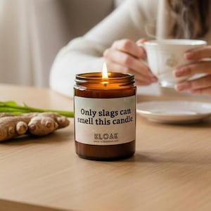 May include: A lit brown glass jar candle with the text "Only slags can smell this candle" and the brand name "KLOAK" on a label. The candle sits on a wooden table next to ginger and celery. A person is in the background.