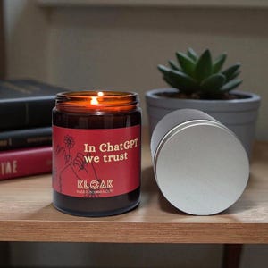 Könnte beinhalten: Eine brennende Kerze in einem Glas mit einem roten Etikett mit der Aufschrift "In ChatGPT we trust". Ein silberner Deckel liegt neben der Kerze. Die Kerze steht auf einer Holzoberfläche mit Büchern und einer kleinen Topfpflanze im Hintergrund.