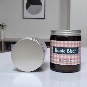 Peut inclure: Un pot de bougie en verre marron avec un couvercle argenté et un couvercle argenté séparé. La bougie a une étiquette à carreaux roses et blancs avec le texte "Basic Bitch" et "KLOAK".