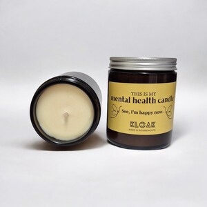 May include: Two glass jar candles with silver lids. One candle is on its side, revealing the wax and wick. The other candle has a label that reads "THIS IS MY mental health candle" and "See, I'm happy now."