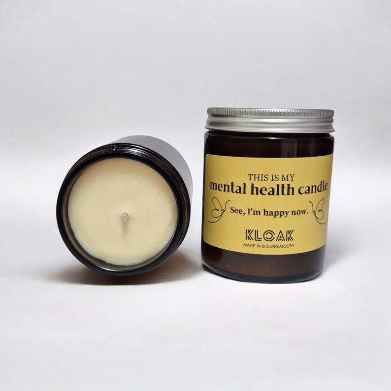May include: Two glass jar candles with silver lids. One candle is on its side, revealing the wax and wick. The other candle has a label that reads "THIS IS MY mental health candle" and "See, I'm happy now."