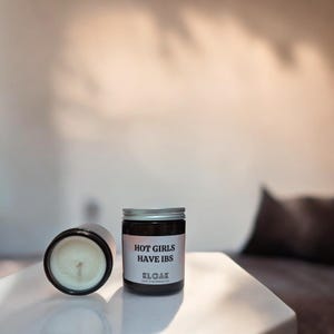 Pode incluir: Um frasco de vela de vidro castanho com uma tampa prateada. A vela tem um r&oacute;tulo branco com o texto "HOT GIRLS HAVE IBS" e o nome da marca "KLOAK" impresso nele.