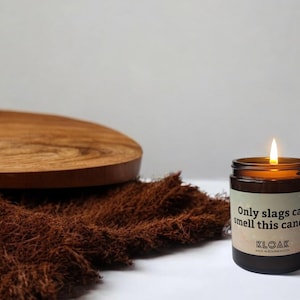 Puede incluir: Una vela encendida en un frasco de vidrio marr&oacute;n con una etiqueta que dice Only slags can smell this candle y KLOAK. La vela tiene una mecha encendida y est&aacute; colocada junto a una tela marr&oacute;n texturizada y una tabla de madera.