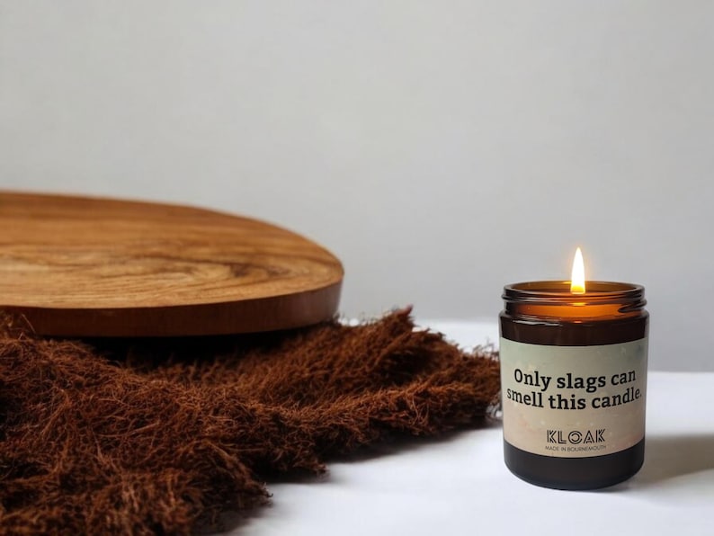 Puede incluir: Una vela encendida en un frasco de vidrio marr&oacute;n con una etiqueta que dice Only slags can smell this candle y KLOAK. La vela tiene una mecha encendida y est&aacute; colocada junto a una tela marr&oacute;n texturizada y una tabla de madera.