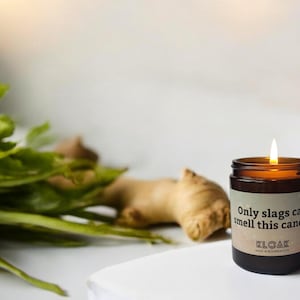Puede incluir: Una vela encendida en un tarro de cristal marr&oacute;n con una etiqueta que dice "Only slags can smell this candle". La vela est&aacute; junto a una ra&iacute;z de jengibre y hojas verdes.