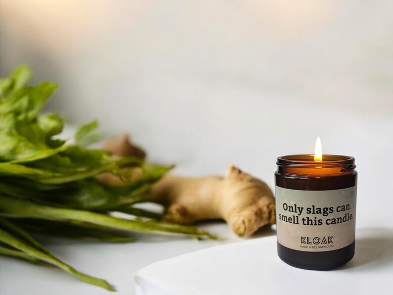 Puede incluir: Una vela encendida en un tarro de cristal marr&oacute;n con una etiqueta que dice "Only slags can smell this candle". La vela est&aacute; junto a una ra&iacute;z de jengibre y hojas verdes.
