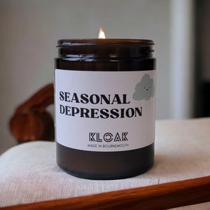 Puede incluir: Una vela encendida en un frasco de vidrio marrón con una etiqueta blanca. La etiqueta dice "SEASONAL DEPRESSION" con un gráfico de nube triste. También se ve el texto "KLOAK MADE IN BOURNEMOUTH". La vela está sobre una superficie blanca.