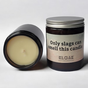 Puede incluir: Dos velas en tarro de vidrio marr&oacute;n oscuro con tapas plateadas. Una vela est&aacute; tumbada de lado, revelando la cera amarilla p&aacute;lida y la mecha. La otra vela tiene una etiqueta que dice "Only slags can smell this candle."