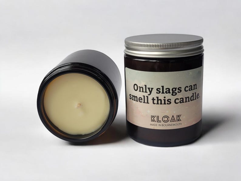 Puede incluir: Dos velas en tarro de vidrio marr&oacute;n oscuro con tapas plateadas. Una vela est&aacute; tumbada de lado, revelando la cera amarilla p&aacute;lida y la mecha. La otra vela tiene una etiqueta que dice "Only slags can smell this candle."